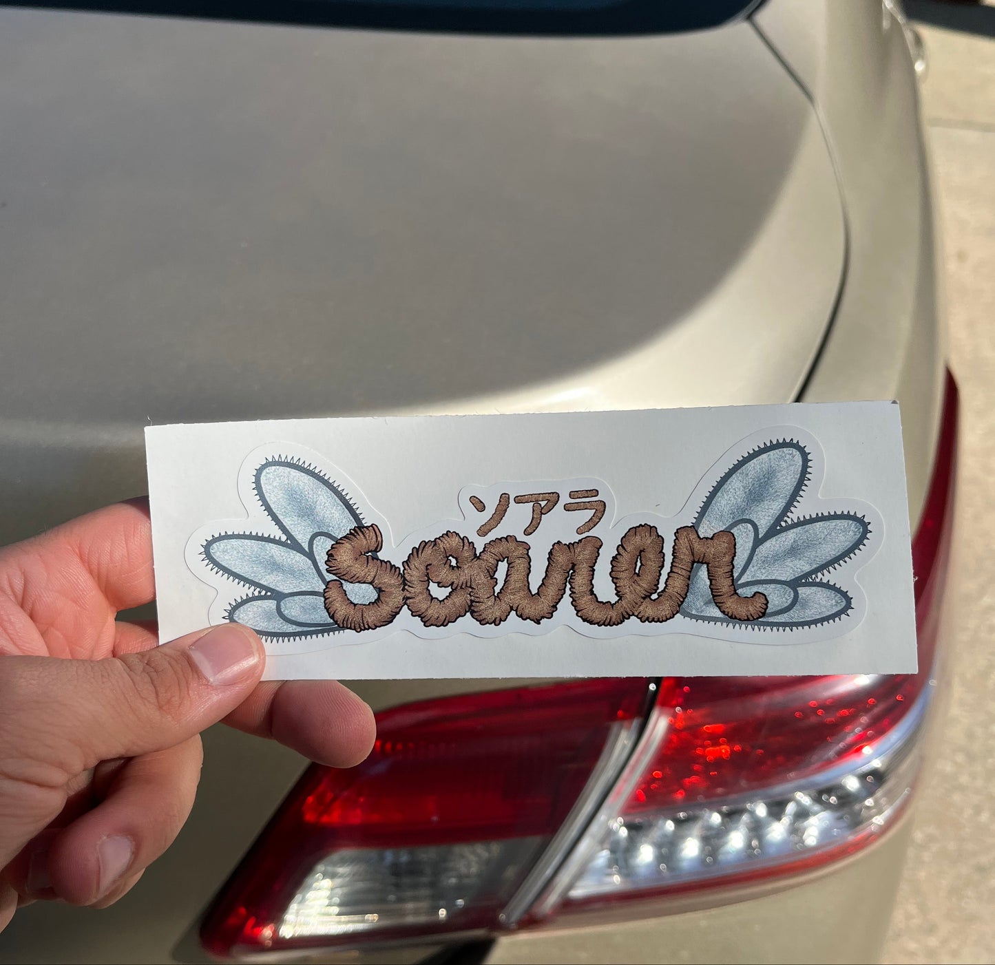 Hand holding a sticker with text and graphics in front of a car's tail light.