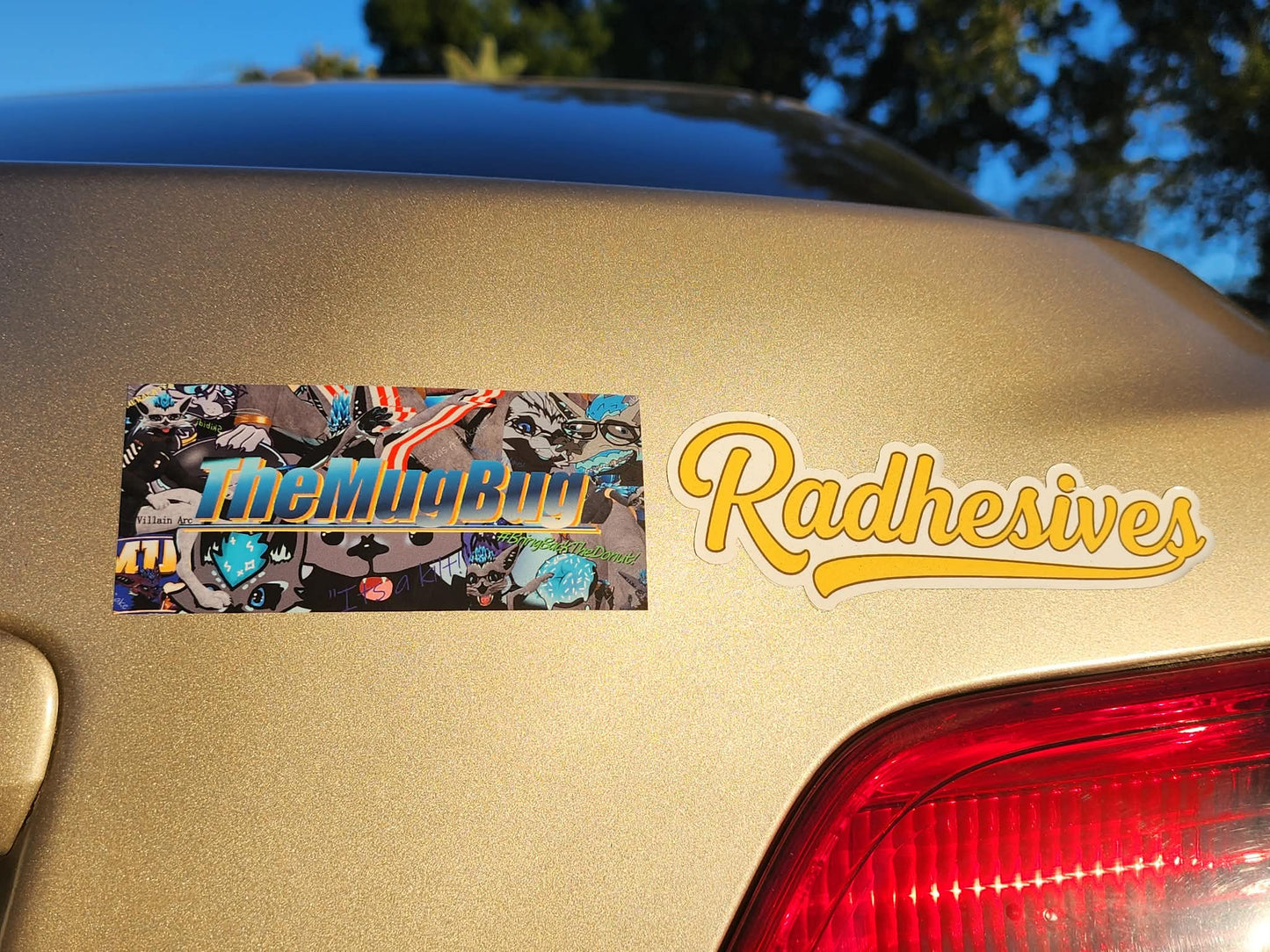 A decal that says "TheMugBug" on it. The decal is on the back of a gold vehicle and next to it is another decal that says "Radhesives".
