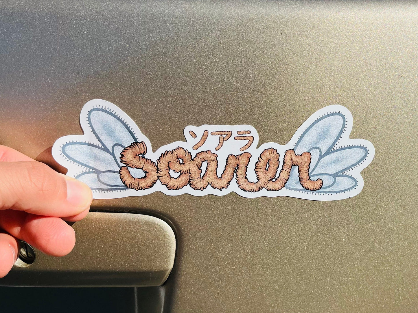 Hand holding a car decal with 'Scener' and 'Radhesive' text on a vehicle's rear window.