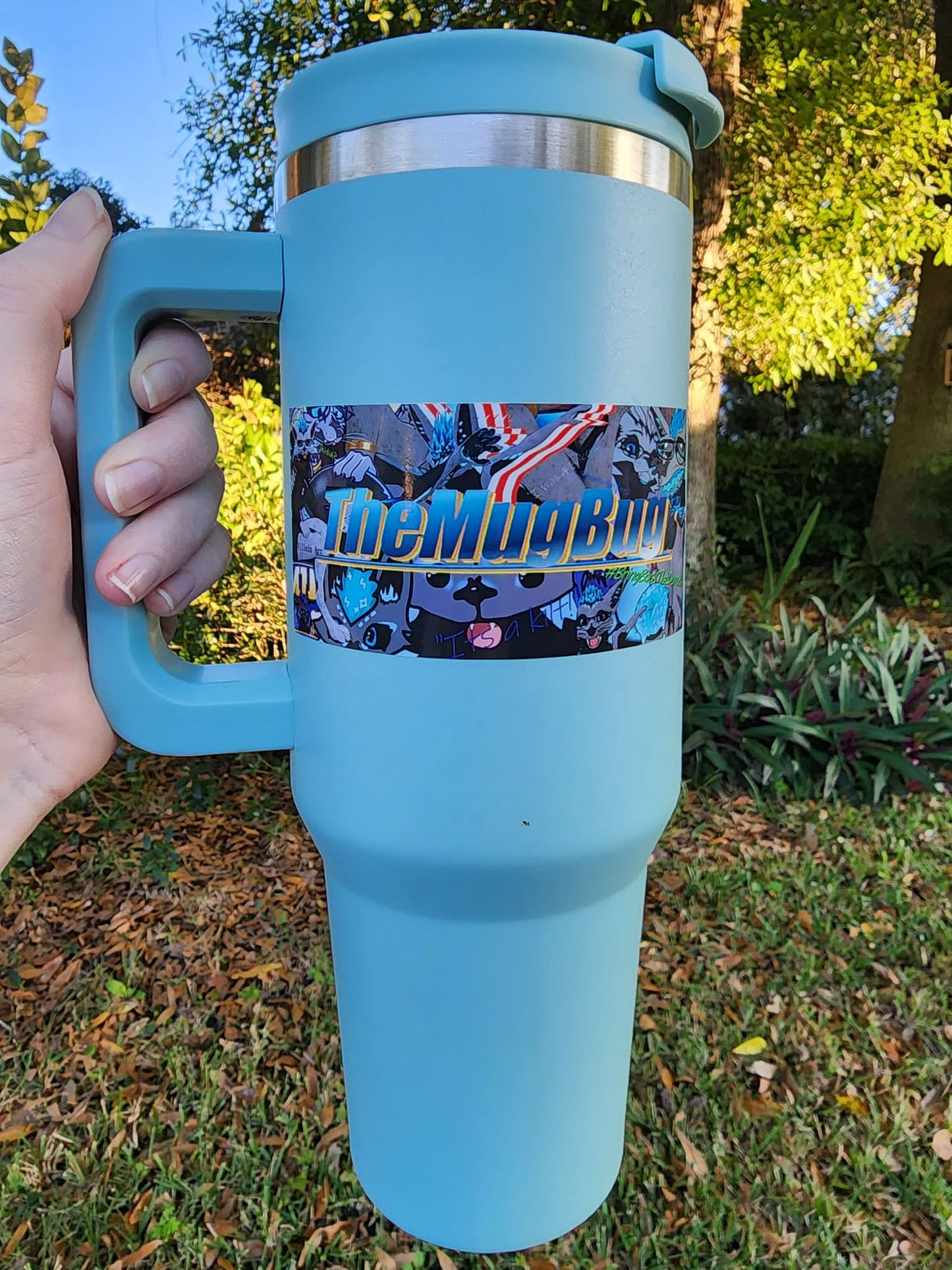 A blue coffee mug with a decal on it that says "TheMugBug". The background is outside with trees, grass, and shrubs.