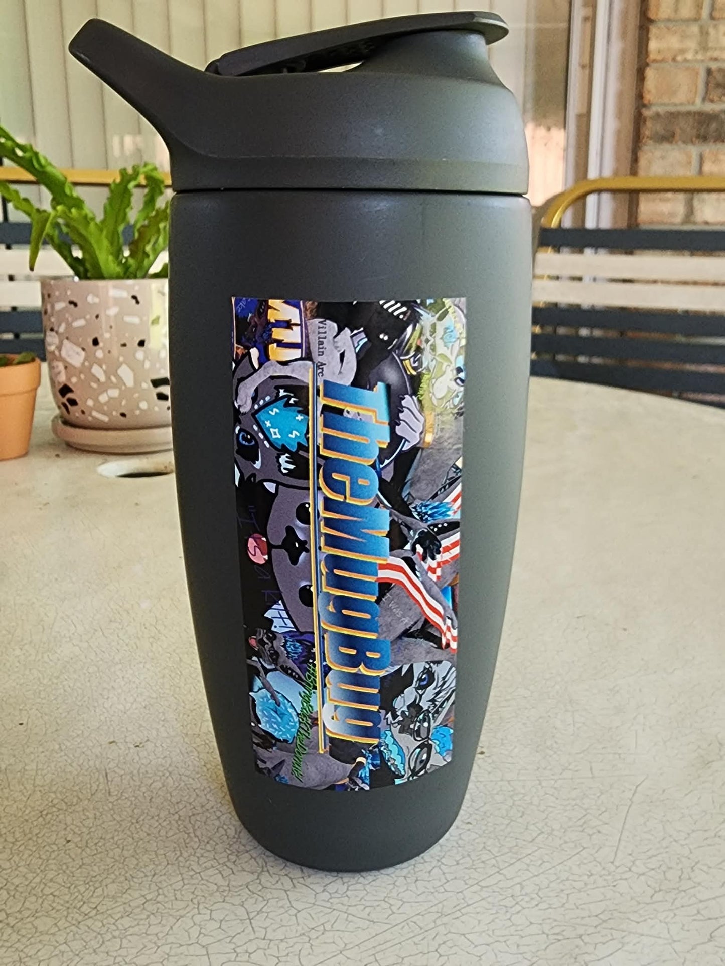 A water bottle with a decal on it set on a table with two houseplants in the background. The decal says "TheMugBug".